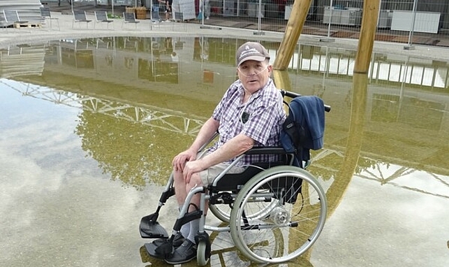 Mann im Rollstuhl genießt kühles Wasser bei der Brunnenskulptur vor dem Festspielhaus