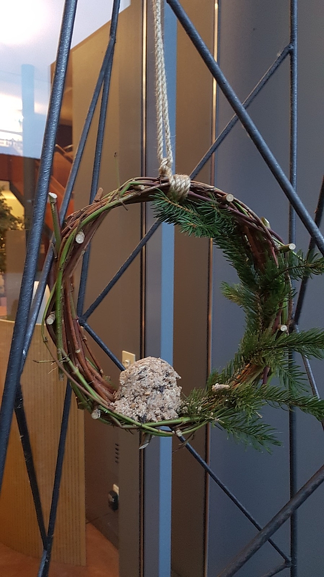 Aus Zweigen geflochten und mit Vogelfutter dekoriert hängt ein fertiger Kranz an einem Gitter.